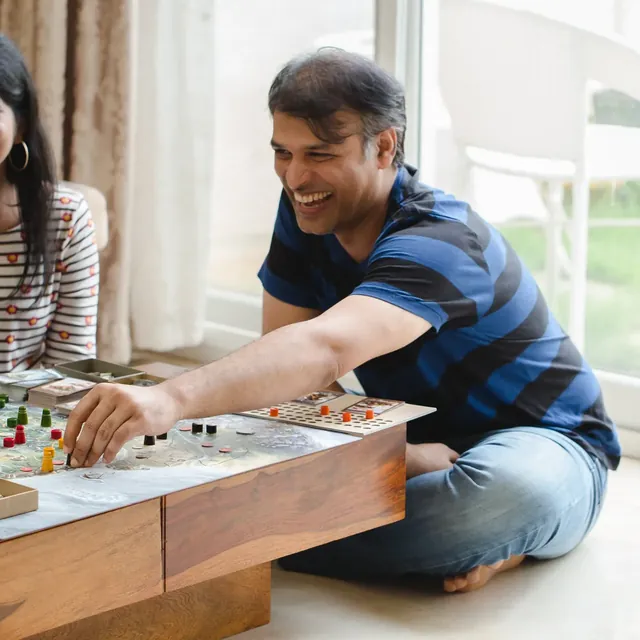 A laughing man in a striped T-shirt sits cross-legged and reaches across the coffee table to move a wooden piece on the map