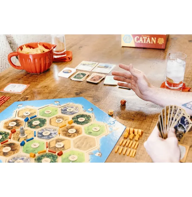 Players trading during a game: one hand reaches out across the table while another holds resource cards, with the board, wooden pieces and dice on a wooden table