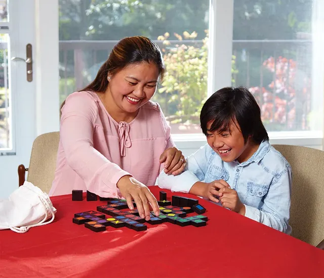 A mother and son smiling as they add tiles to a Qwirkle layout on a red table