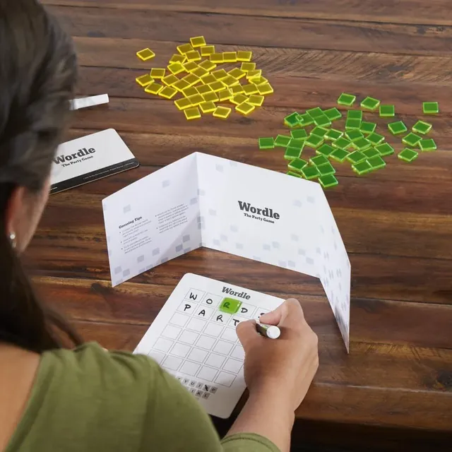 A player writing a guess on a Wordle board behind a shield, with green and yellow tiles on the table