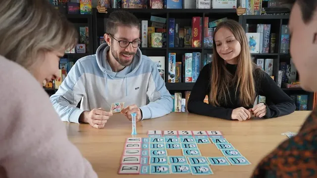 Three adults playing at a table, with a grid of cards and a sand timer