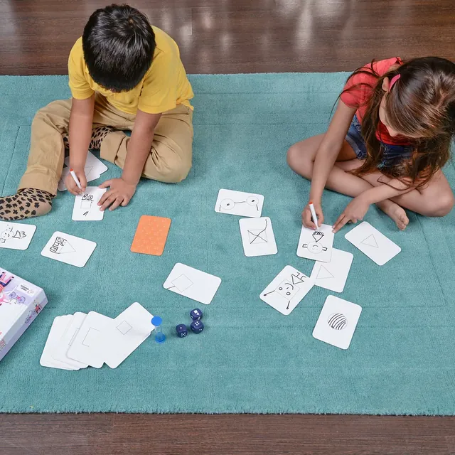A top view of two children drawing on cards on a rug, with the dice and box beside them
