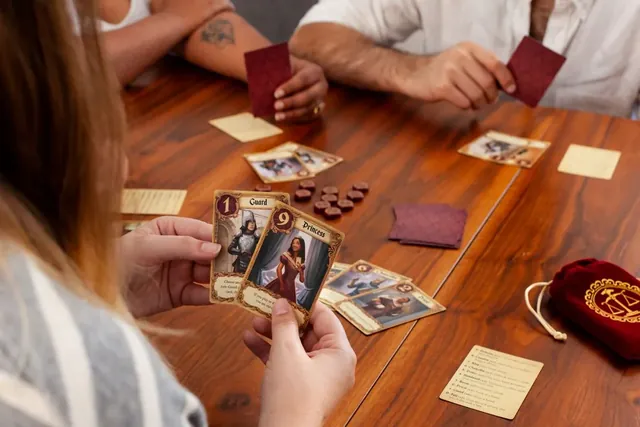 Players holding Love Letter cards at a wooden table, with favour tokens and the cloth bag