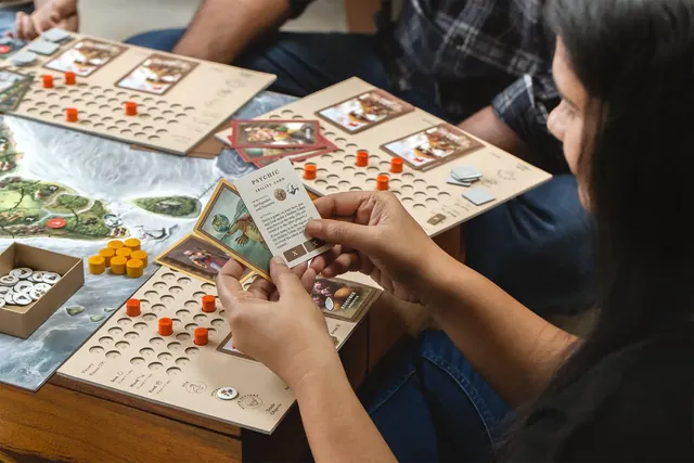 A player holds the Psychic ability card above her tracker board, which has orange wooden pegs and two army cards on it, with other players' boards and the map beside her