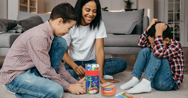 A mother and two boys playing on a living-room floor, one boy holding his head as he thinks