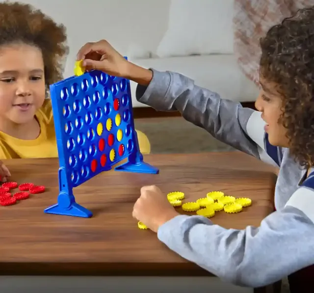 Two children playing at a wooden table: one reaches over to drop a yellow disc into the blue grid while the other watches, with a pile of red discs on one side and yellow discs on the other
