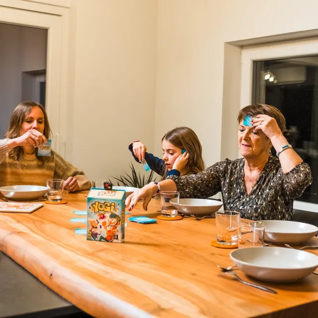 A family playing round a dinner table, one with a card on her forehead