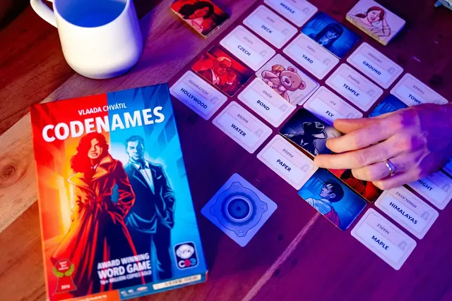 A hand pointing at a word card in the grid, next to the box and a coffee mug
