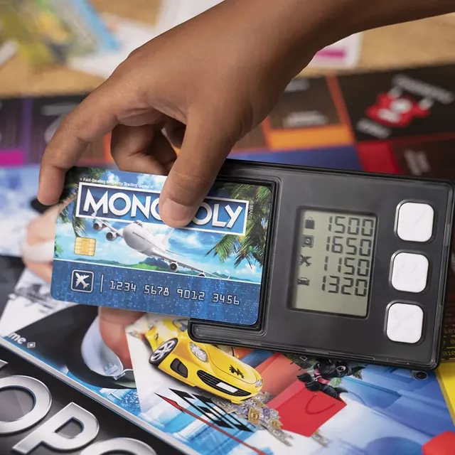A hand tapping a Monopoly bank card on the banking unit, which shows the players' balances