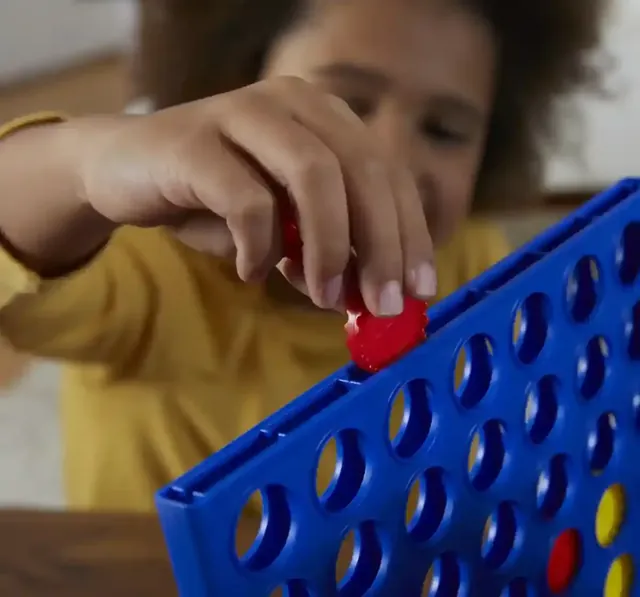Close-up of a child's hand pushing a red ridged disc into a slot at the top of the blue grid, with the child's face blurred in the background
