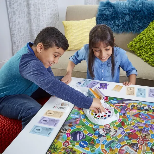 A boy spins the spinner while a girl watches, with cards and money around the board