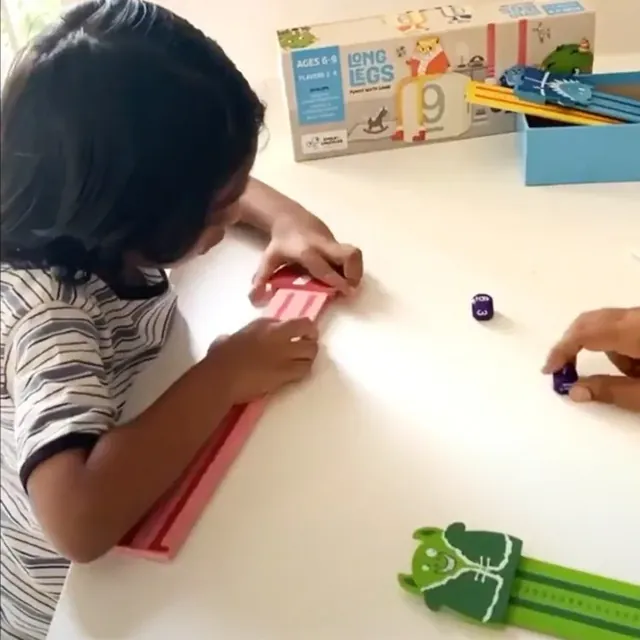 A child seen from the side, sliding a monster along a ruler on a table, with the box behind