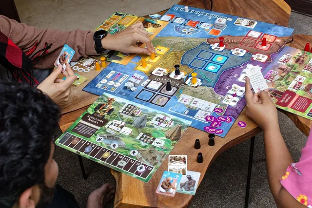 Players round a wooden table with the board and their cards, seen from above