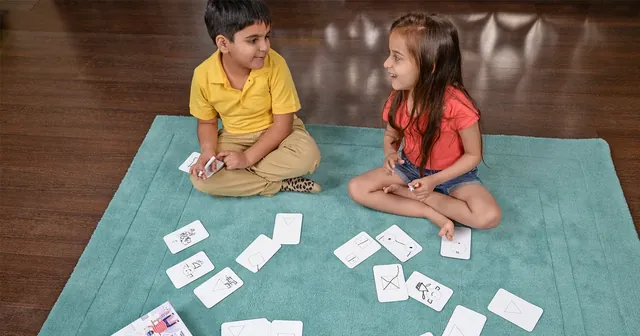 Two children sitting on a rug with drawn cards between them, seen from a distance